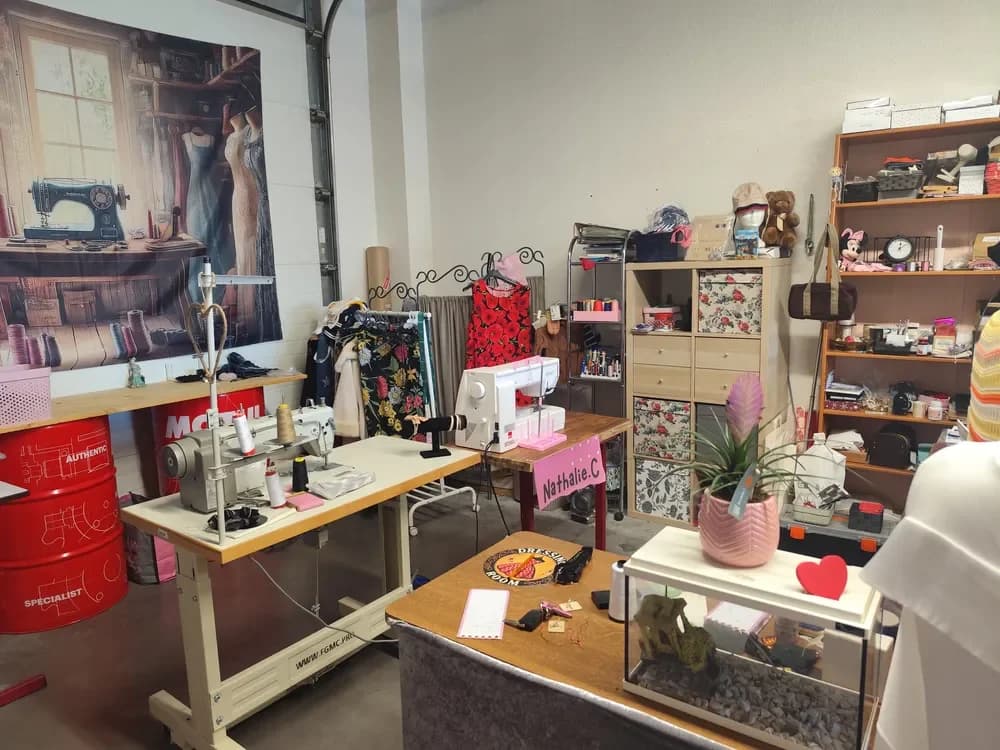 L'atelier CÉKIKAFÉ : au premier plan, une machine à coudre industrielle et une machine à coudre blanche sur leurs tables de travail, avec des cônes de fil. Derrière, un portant de robes à fleurs rouges et noires, un meuble à cases avec des boîtes en tissu fleuri, des étagères remplies de fournitures, de peluches et de boîtes de rangement. Au mur, une grande affiche représentant une machine à coudre ancienne devant des robes
