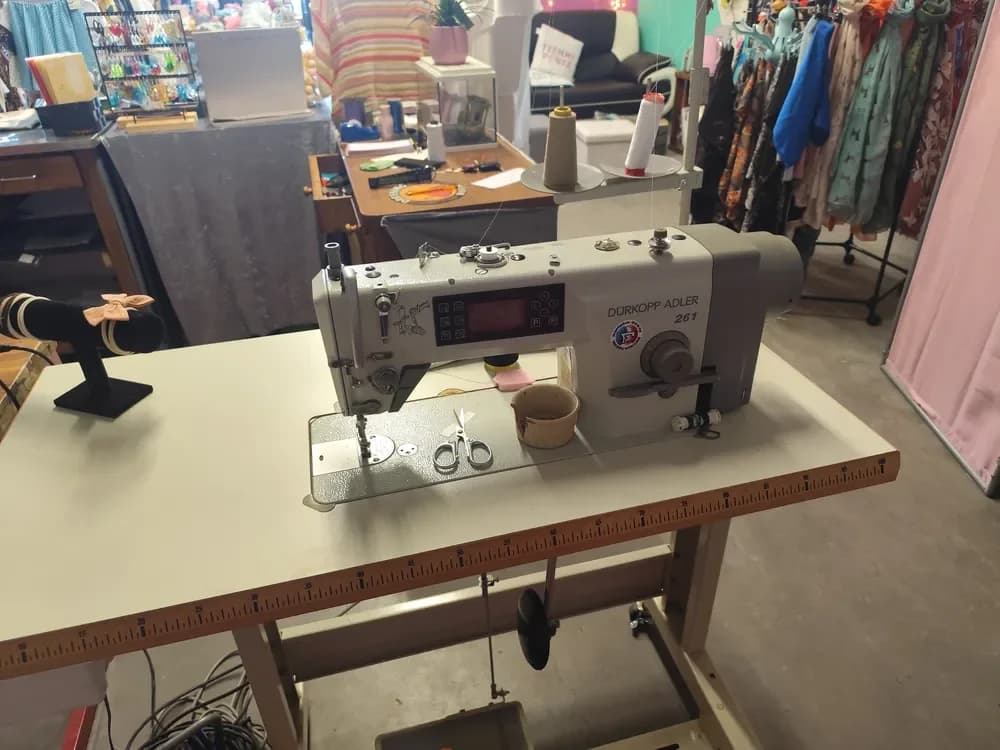 La machine à coudre industrielle Dürkopp Adler 261 de l'atelier, grise avec un écran de commande, installée sur une grande table blanche bordée d'un mètre ruban, avec une paire de ciseaux posée sur le plateau et deux cônes de fil au-dessus. En arrière-plan, la boutique avec un portant de foulards colorés et un présentoir de bijoux