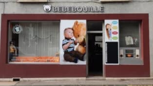 Façade de l'ancienne boutique BébéBouille, encadrée de bordeaux, avec l'enseigne « BEBEBOUILLE » et sa tête d'ourson en lettres blanches. Dans la grande vitrine, une affiche montre un bébé assis contre un gros ours en peluche, à côté de vêtements pour bébé exposés. À droite, une porte vitrée et une petite vitrine avec une affiche et des articles