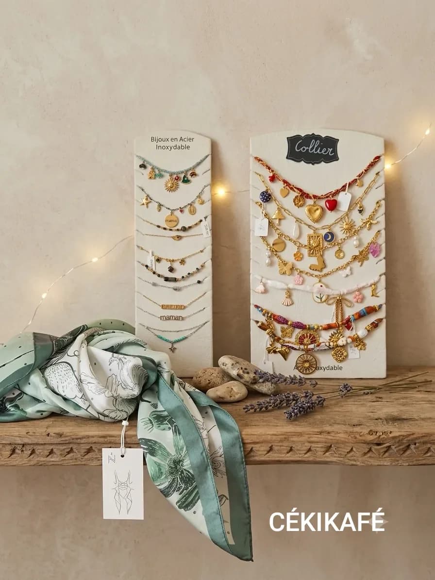Deux présentoirs blancs posés sur une étagère en bois ancien, devant un mur beige éclairé d'une guirlande lumineuse. À gauche, un présentoir « Bijoux en acier inoxydable » garni de fins bracelets dorés à breloques, dont des médailles « mamie » et « maman ». À droite, un présentoir « Collier » garni de colliers dorés à breloques colorées : cœurs rouges, soleils, papillons et perles. Au premier plan, un foulard en soie vert d'eau à motifs floraux, des galets et des brins de lavande
