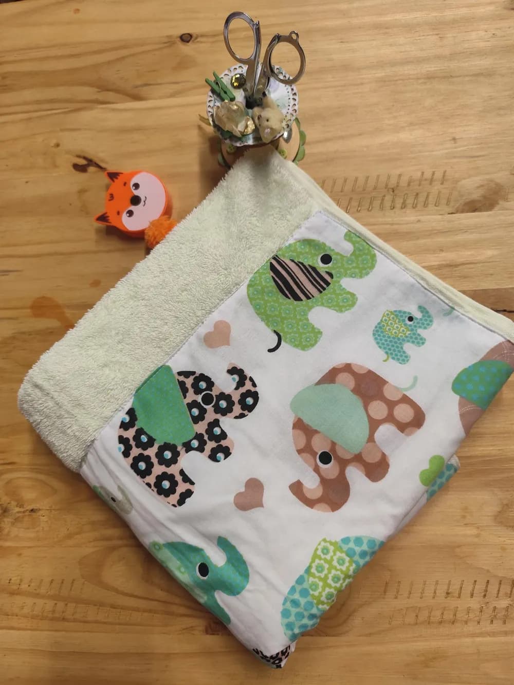 Linge en éponge vert très pâle, plié sur une table en bois, avec une large bande de coton blanc cousue et imprimée d'éléphants à motifs verts, turquoise, marron et roses, et de petits cœurs