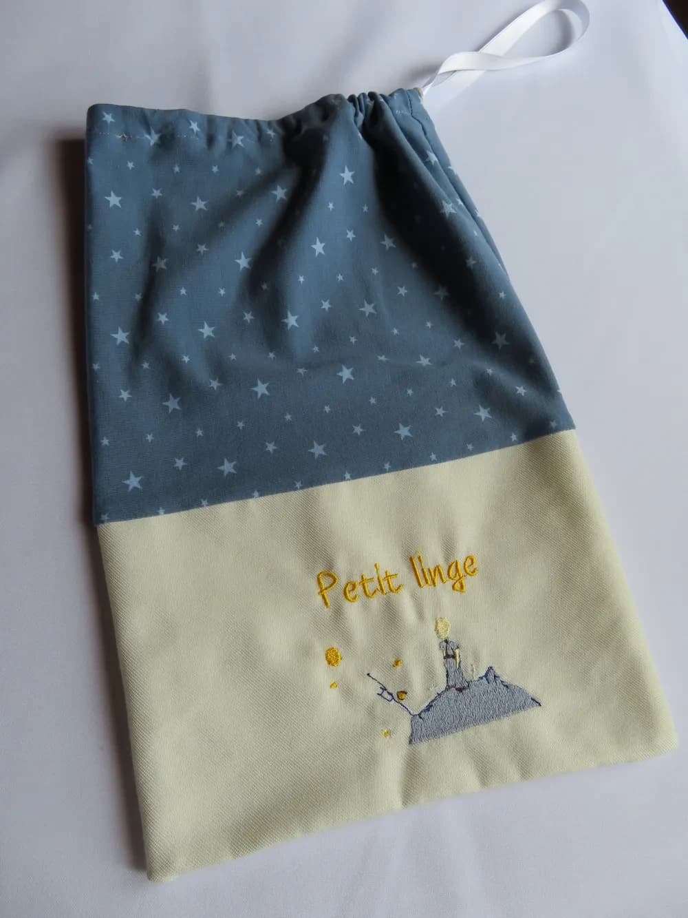 Sac pochon pour enfant posé à plat sur un fond blanc, fermé en haut par un cordon froncé et une boucle de ruban blanc. La partie haute est en tissu bleu-gris semé de petites étoiles blanches, la partie basse en tissu jaune très pâle, brodée en fil jaune des mots « Petit linge » et d'un petit personnage aux cheveux blonds, debout sur une planète grise, entouré de petites étoiles dorées