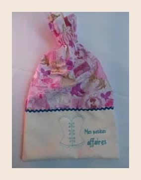 Sac pochon vu en entier, debout et fermé par son cordon froncé en haut. La partie supérieure est en tissu imprimé Mademoiselle Paris rose et violet, séparée par un galon ondulé bleu canard d'une large bande en tissu écru brodée en fil turquoise d'un corset et des mots « Mes petites affaires »