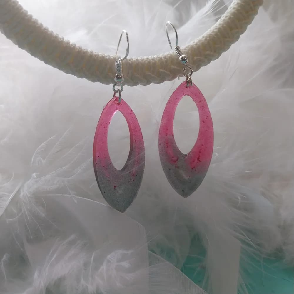 Boucles d'oreilles marquise en résine dégradée rose et gris, suspendues à un support en corde beige sur fond de plumes blanches