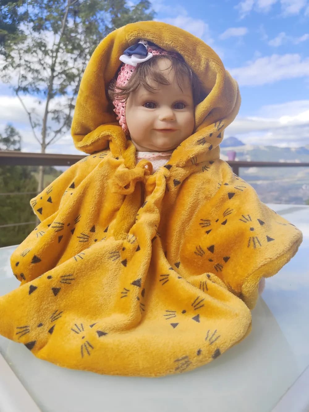 Poupée bébé assise sur une table en verre en extérieur, enveloppée dans une cape polaire jaune moutarde à capuche, imprimée de petites têtes de chat noires et fermée par un lien noué, avec des montagnes et un ciel bleu en arrière-plan