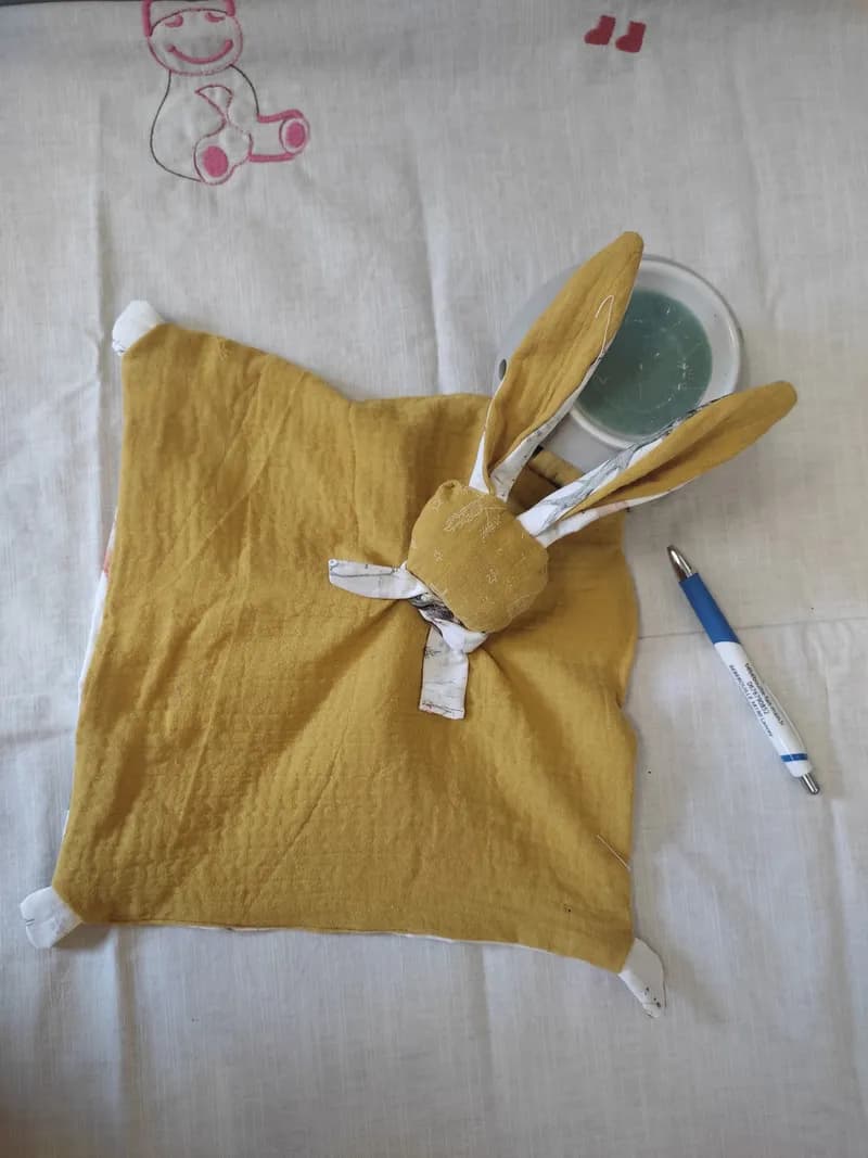 Doudou plat en forme de lapin en gaze de coton moutarde avec de longues oreilles, posé sur un lange en tissu brodé