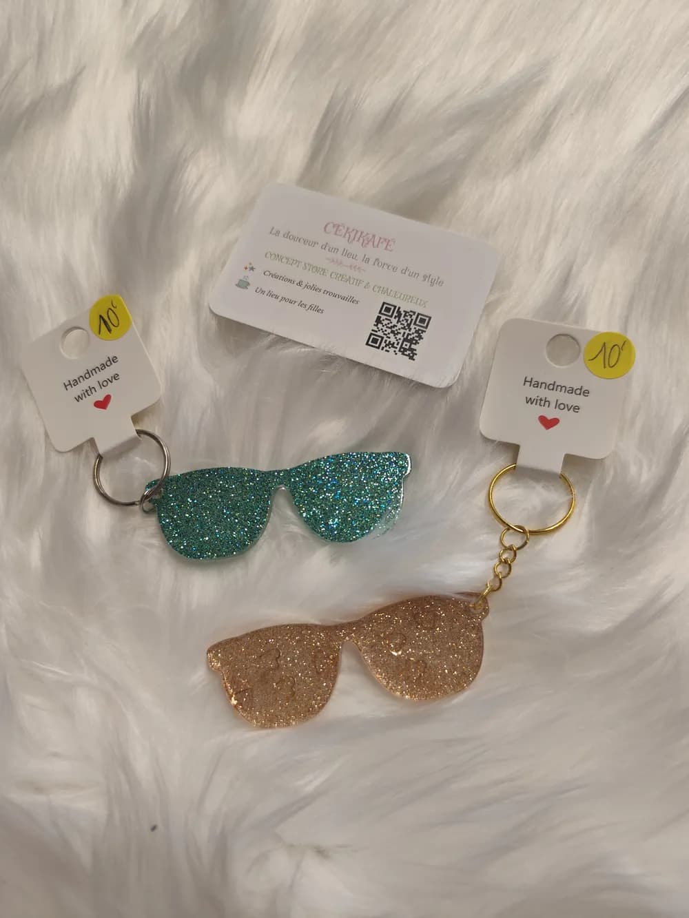 Deux porte-clés en résine pailletée en forme de lunettes de soleil, l'un turquoise et l'autre doré, sur une étiquette « Handmade with love »