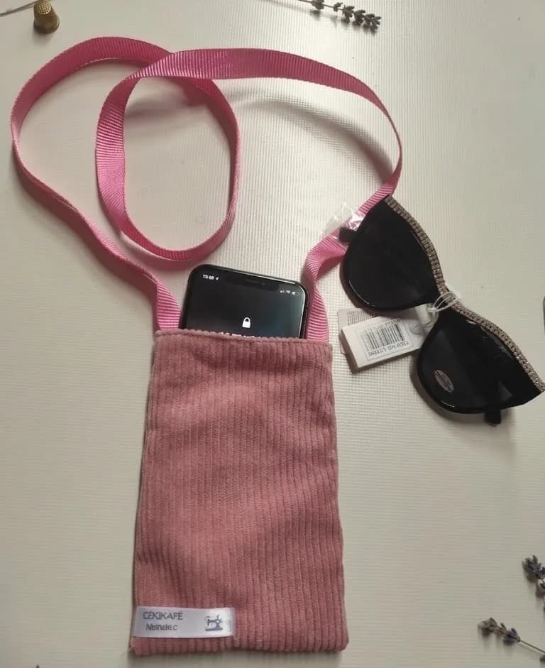 Pochette téléphone en velours côtelé rose avec cordon bandoulière rose, contenant un smartphone, posée à côté de lunettes de soleil