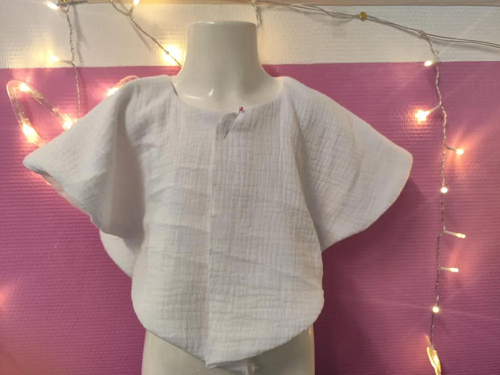 Cape de baptême en gaze de coton blanche portée sur un buste mannequin, manches courtes évasées en forme d'ailes, fermeture par une épingle au centre devant, broderie pas encore réalisée sur cette photo qui illustre la coupe de la cape, fond rose avec guirlande lumineuse