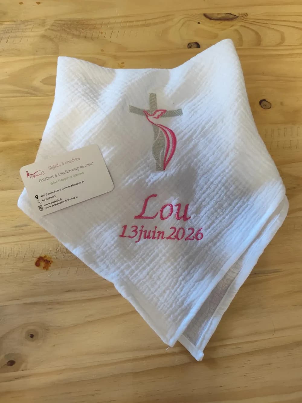 Carré de baptême chrémeau en gaze de coton blanche plié en triangle, posé sur une table en bois, brodé d'une croix gris taupe avec une colombe rose en son centre, ainsi que du prénom Lou et de la date 13 juin 2026 en fil rose, avec la carte de visite CÉKIKAFÉ posée à côté