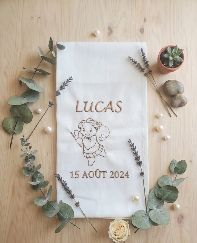 Écharpe de baptême en tissu écru posée à plat sur une table en bois, brodée en fil doré du prénom LUCAS, d'un ange musicien tenant un fin instrument et de la date 15 AOÛT 2024, entourée de branches d'eucalyptus, de brins de lavande, de perles, d'une rose et d'un petit pot de succulente en décor, exemple de réalisation avec un autre prénom que celui choisi à la commande