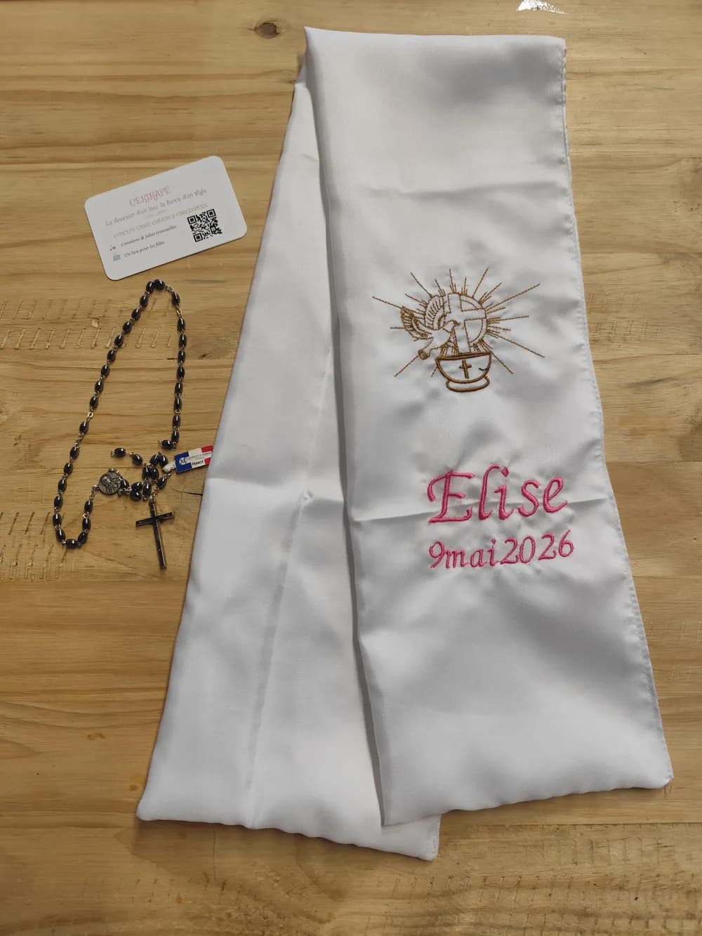 Écharpe de baptême en tissu blanc satiné pliée sur une table en bois, brodée d'un motif calice doré surmonté d'une croix, entouré de rayons de soleil et d'une colombe, avec le prénom Elise et la date 9 mai 2026 brodés en fil rose en dessous, un chapelet à grains noirs et croix posé à côté en décor, exemple de réalisation avec un autre prénom que celui choisi à la commande