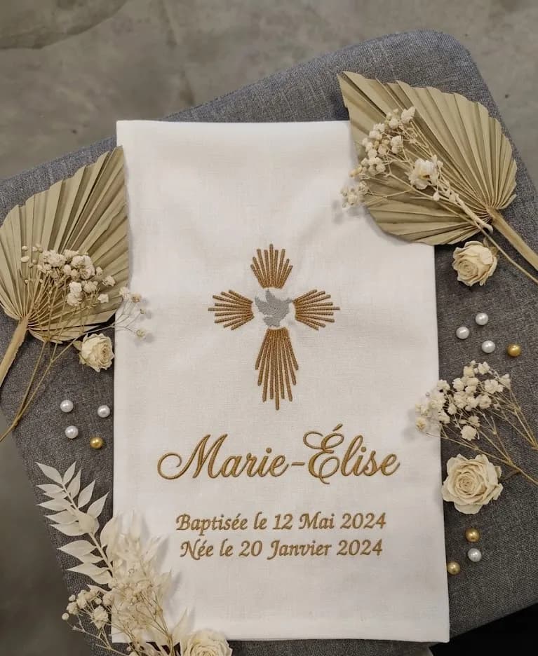Écharpe de baptême en coton blanc posée sur un tissu gris, brodée d'une croix moderne dorée à rayons avec une colombe grise en son centre, ainsi que du prénom Marie-Élise et des dates « Baptisée le 12 Mai 2024 » et « Née le 20 Janvier 2024 » en fil doré, entourée de palmes séchées, de fleurs séchées blanches et de perles dorées et blanches, exemple de réalisation avec un autre prénom que celui choisi à la commande