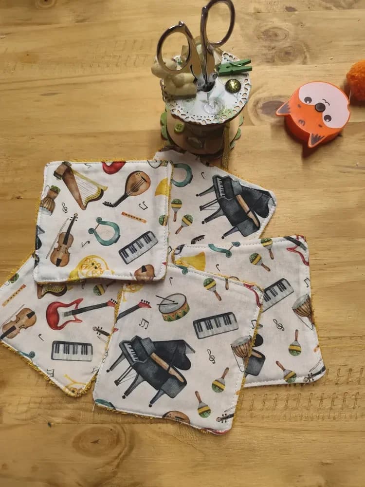 Cinq lingettes lavables carrées étalées sur une table en bois, côté coton blanc cassé imprimé d'instruments de musique : pianos à queue noirs, claviers, violon, harpe, lyre turquoise, mandoline, guitare électrique rouge, cor doré, tambour, maracas et notes. En haut, une bobine de couture décorée avec des ciseaux et un mètre ruban orange en forme de tête de renard.