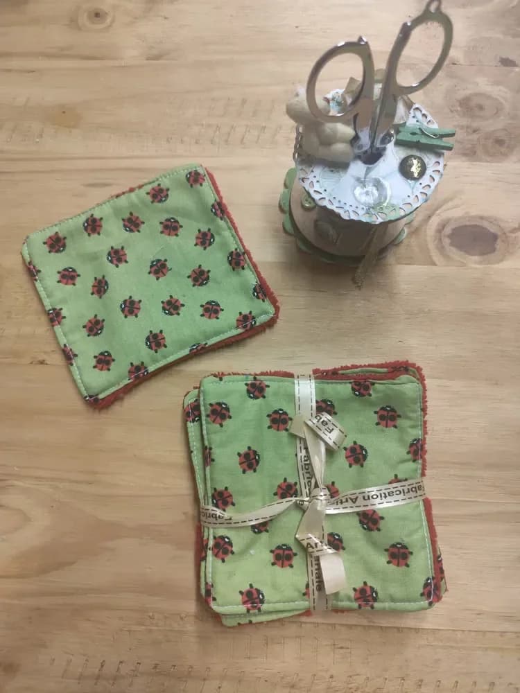 Lingettes lavables vertes à coccinelles rouges et noires, éponge de bambou rouge ou bleu turquoise – CÉKIKAFÉ. Une lingette posée à plat et un lot de lingettes noué d'un ruban doré « Fabrication Artisanale », côté coton vert pomme imprimé de petites coccinelles, sur une table en bois sous une bobine de couture décorée et des ciseaux.