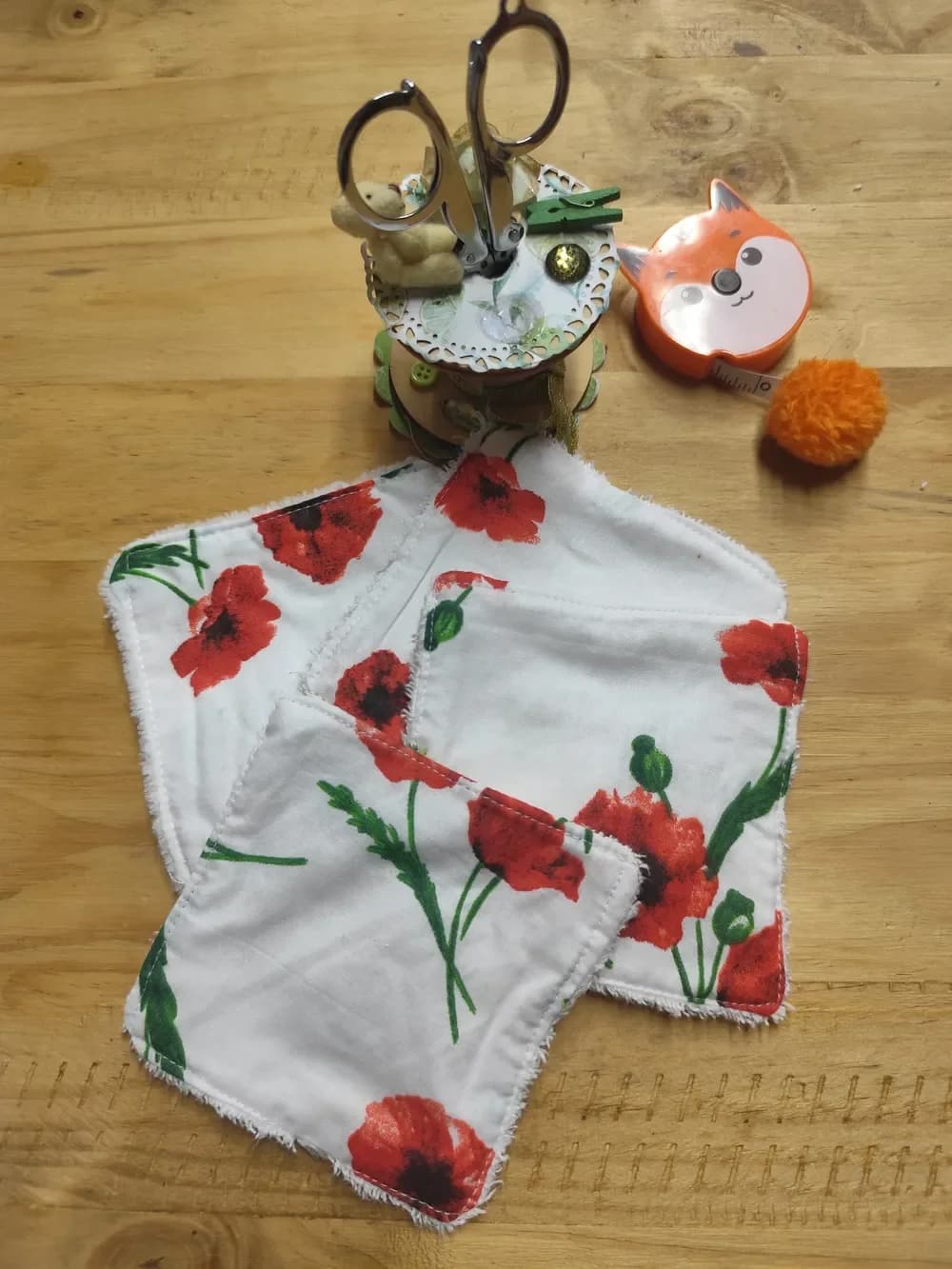 Lingettes démaquillantes lavables carrées, côté coton blanc imprimé de grands coquelicots rouges au cœur noir, avec leurs tiges et boutons verts, étalées en éventail sur une table en bois clair. On aperçoit sur les bords la bordure en éponge blanche. En arrière-plan, un présentoir de couture rond avec une paire de ciseaux argentés, un petit ourson et une pince à linge verte, ainsi qu'un mètre ruban orange en forme de tête de renard et un pompon orange.