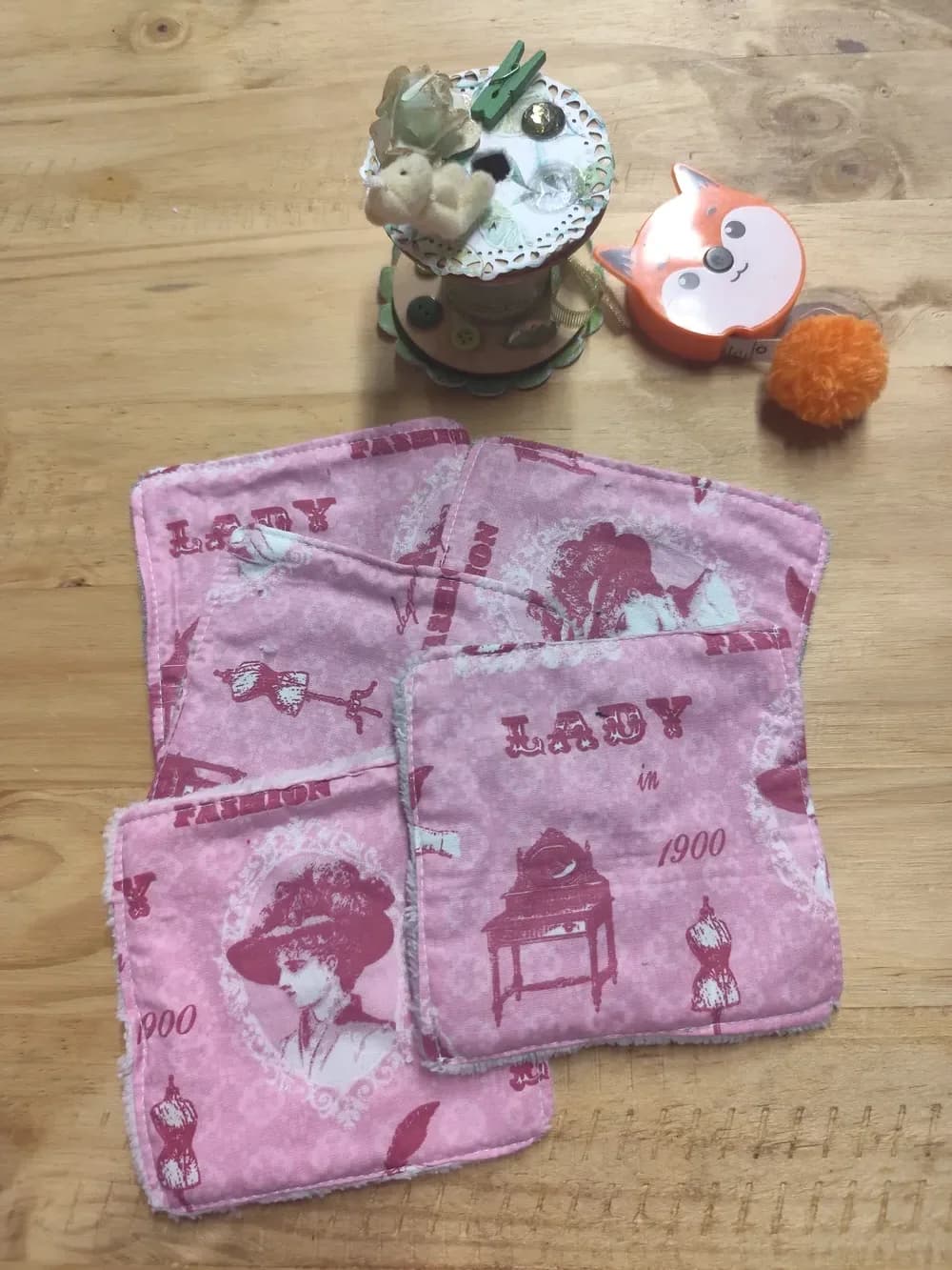 Lingettes lavables roses Lady 1900, silhouette de femme en bustier et chapeau, éponge de bambou rose et grise – CÉKIKAFÉ. Les cinq lingettes sont étalées côté imprimé sur une table en bois : tissu rose aux motifs bordeaux, avec le portrait d'une femme au grand chapeau dans un médaillon, des bustiers de couturière, une coiffeuse ancienne et les mots « LADY in 1900 » et « FASHION ». En arrière-plan, une bobine de couture décorée d'un petit ourson et un mètre ruban orange en forme de tête de renard.