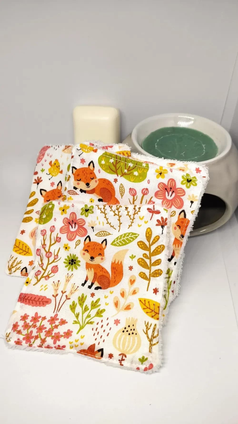 Lingettes lavables en coton bio certifié GOTS, motifs renards, fleurs et arbres, éponge au choix – CÉKIKAFÉ. Gros plan de trois lingettes dressées sur fond blanc, côté coton blanc imprimé d'un petit renard orange assis qui sourit, d'un renard endormi, de fleurs roses et corail, de feuilles moutarde et vertes et d'un poussin jaune, avec la bordure en éponge blanche. Derrière, un savon blanc et un brûle-parfum en céramique grise.