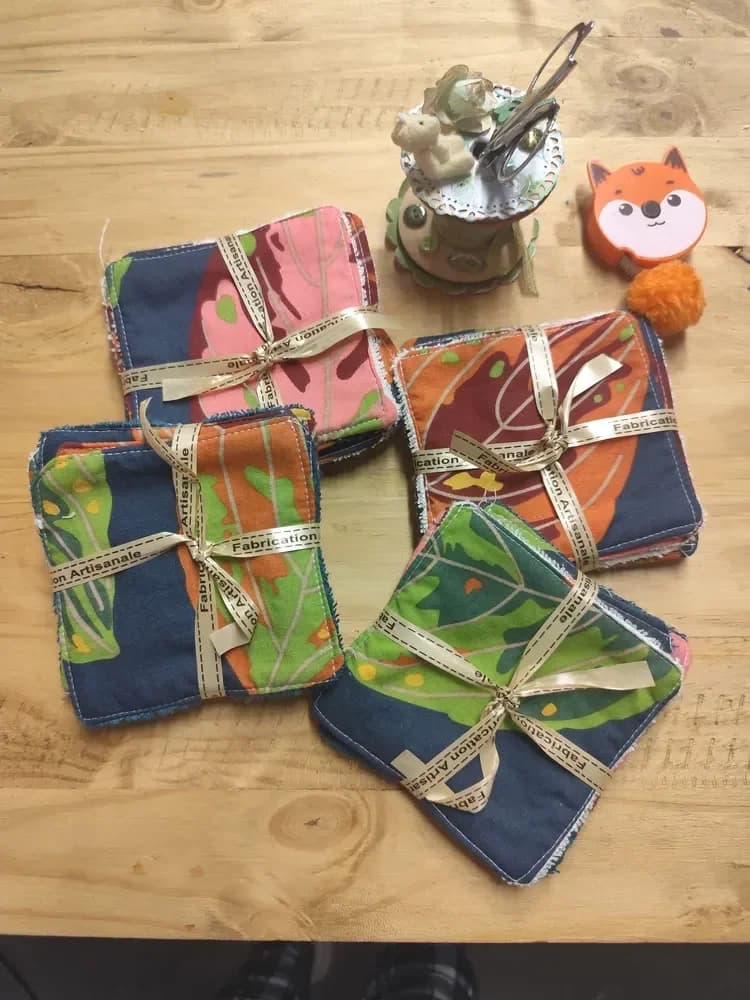Lingettes surprise lavables en coton coloré, formes vertes, bleues, jaunes, roses et orange, éponge de bambou – CÉKIKAFÉ. Quatre lots de lingettes empilées, chacun noué d'un ruban doré « Fabrication Artisanale », sur une table en bois : le coton bleu marine est imprimé de grandes feuilles stylisées vertes, orange, roses et bordeaux, différentes d'un lot à l'autre. En haut, une bobine de couture décorée avec des ciseaux et un mètre ruban orange en forme de tête de renard.