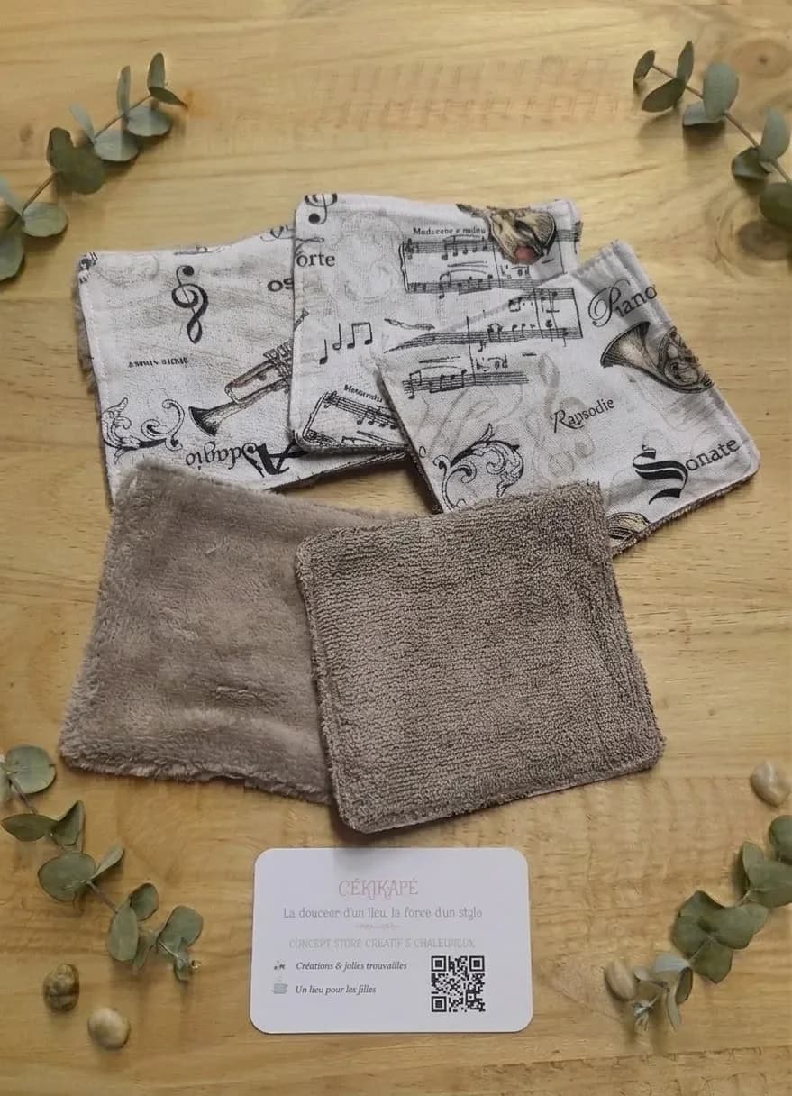 Cinq lingettes lavables carrées posées sur une table en bois. Trois montrent la face en coton écru imprimé de partitions, de notes de musique, d'une clé de sol, d'une trompette et d'un cor, avec les mots « Adagio », « Forte », « Piano », « Rapsodie » et « Sonate ». Les deux autres montrent la face en éponge de bambou beige foncé. Autour, des brins d'eucalyptus, des galets et une carte de visite CÉKIKAFÉ.