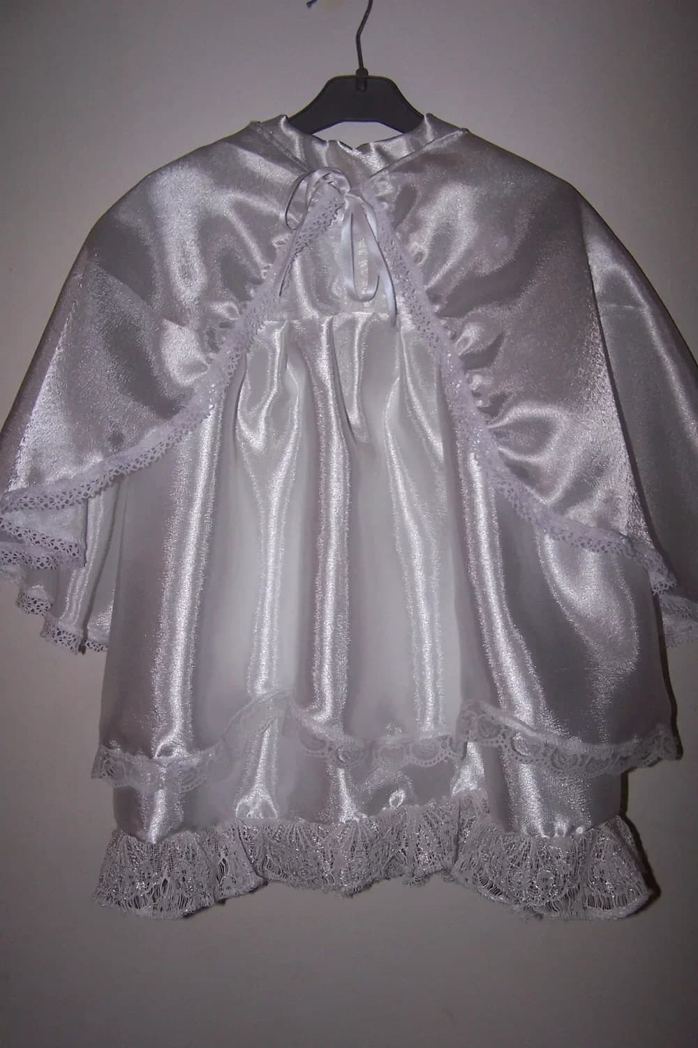 Robe de baptême blanche CÉKIKAFÉ et sa cape présentées sur un cintre noir devant un mur clair, vues de face : la cape en satin blanc brillant, nouée au cou par un ruban de satin blanc et bordée d'une dentelle blanche ajourée, retombe sur les épaules et s'ouvre sur le devant de la robe en satin blanc, qui se termine par un double volant bordé de dentelle blanche.