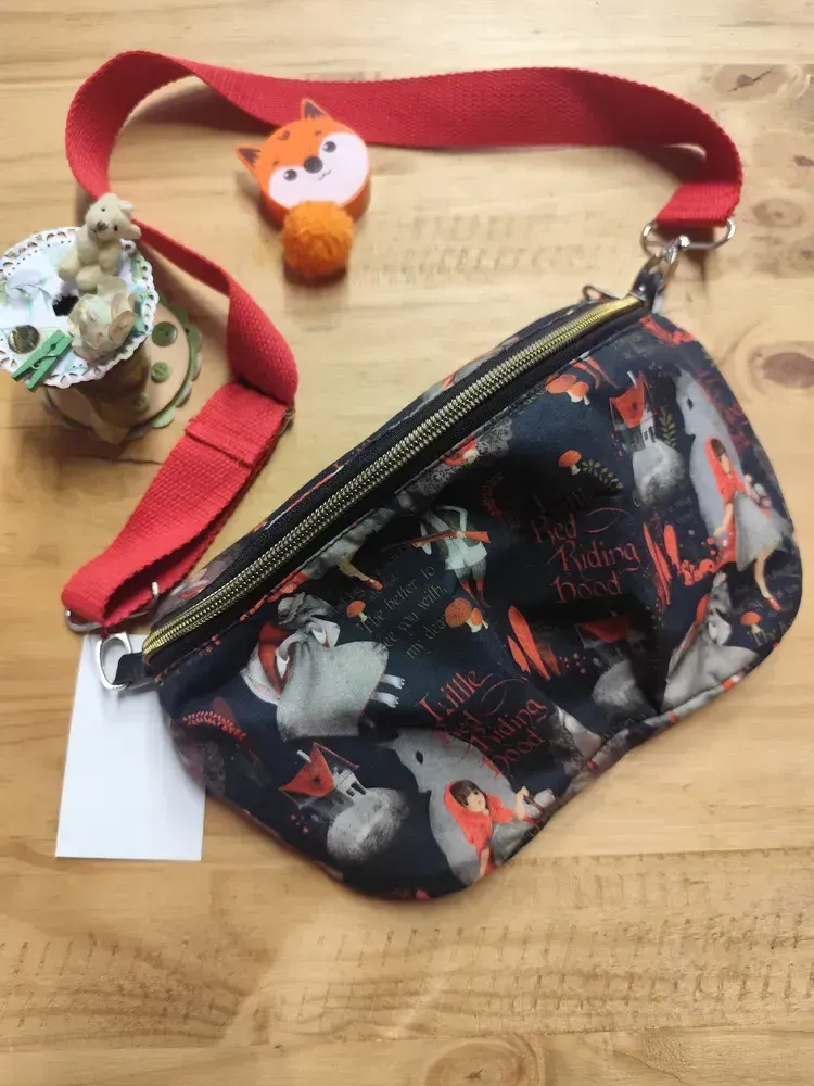 Le sac banane posé de biais sur une table en bois clair, fermeture éclair dorée en diagonale, avec son anse rouge en sangle de coton entière, réglable par une boucle et accrochée de chaque côté par un mousqueton argenté. Le tissu noir montre le Petit Chaperon rouge, le loup, la grand-mère et l'inscription « Little Red Riding Hood ». Autour, un mètre ruban renard orange à pompon, une bobine décorée d'un petit ours et une étiquette blanche.