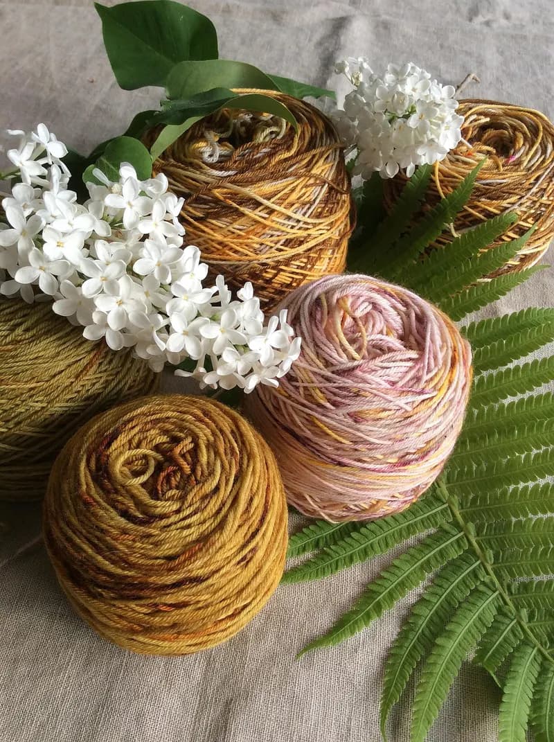 Cinq pelotes de fil naturel aux teintes moutarde, caramel, vert olive et rose pâle chiné, posées sur une toile de lin beige, entourées de brins de lilas blanc et de feuilles de fougère vertes