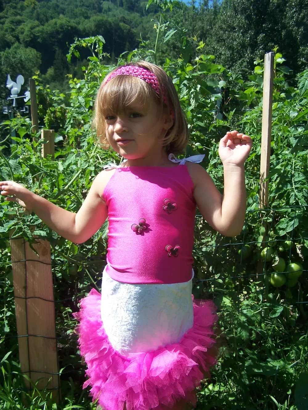 Petite fille dans un potager, un bras levé, en robe tutu au haut rose fuchsia orné de petites fleurs, avec une ceinture de dentelle blanche et une jupe en tulle rose fuchsia très bouffante, un bandeau rose dans les cheveux