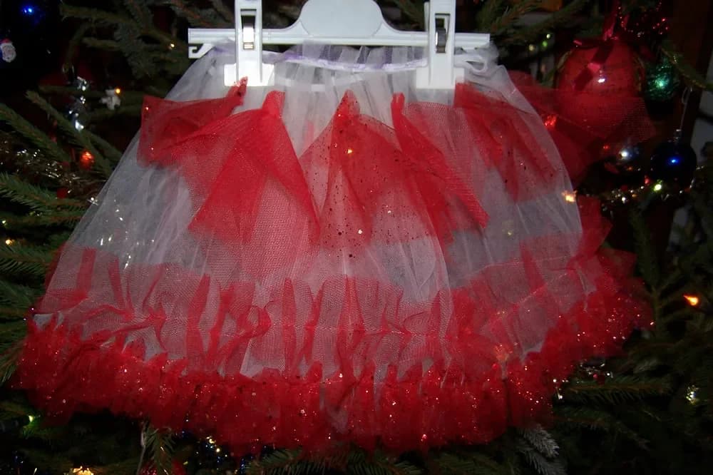 Jupe tutu suspendue à un cintre blanc devant un sapin de Noël illuminé, faite de couches superposées de tulle rouge pailleté et de tulle blanc