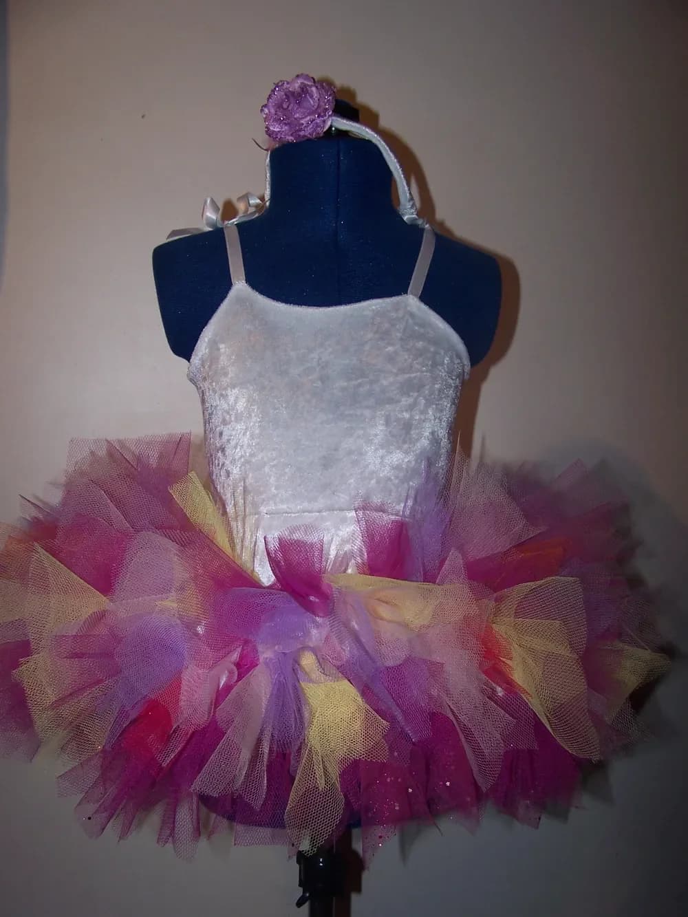 Robe tutu présentée sur un buste de couture bleu marine : haut en tissu blanc brillant à fines bretelles et jupe en tulle très fournie mêlant le fuchsia, le violet, le rose et le jaune, avec une fleur violette posée en haut du buste
