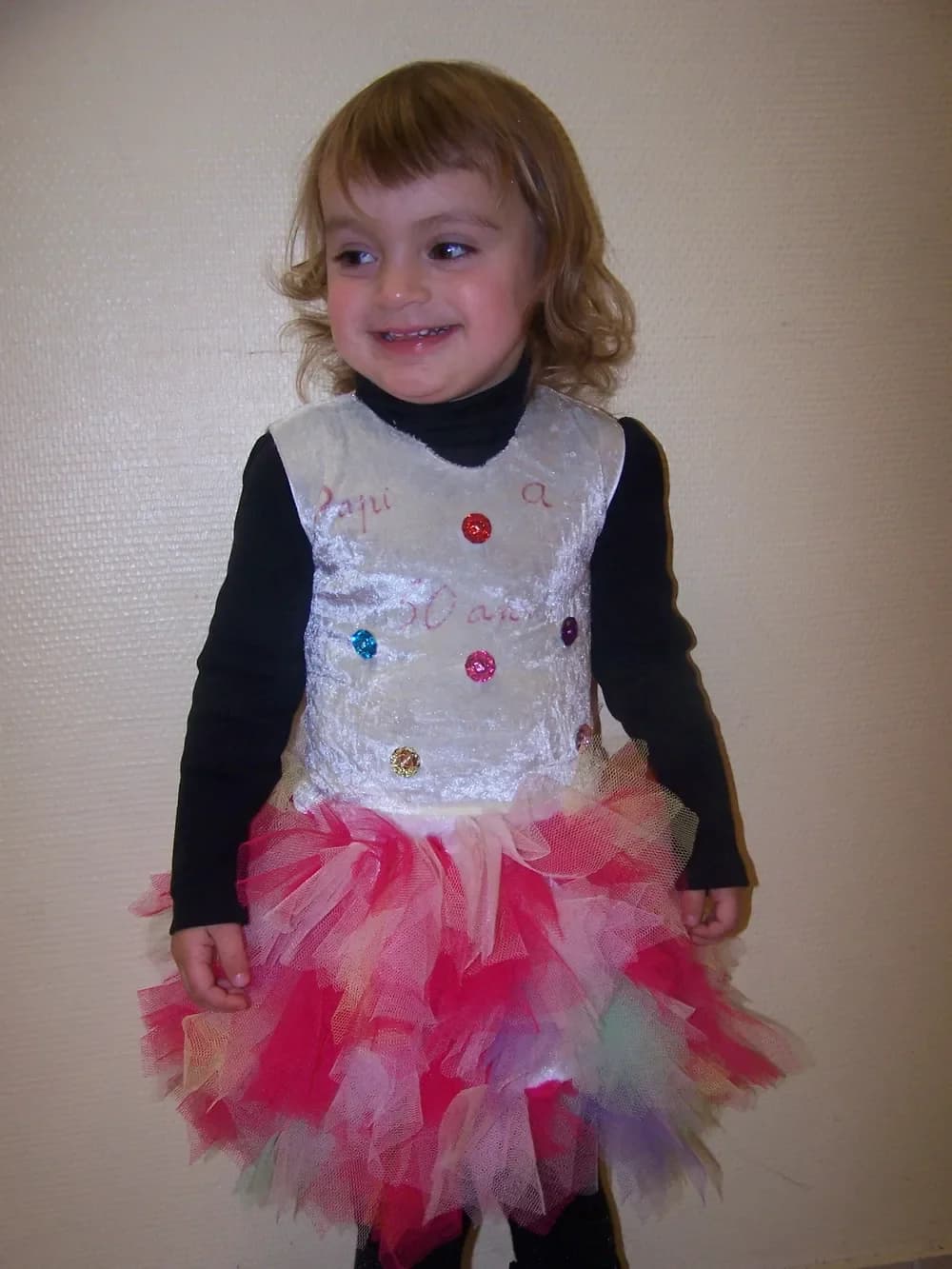 Petite fille souriante portant un pull noir à manches longues sous une robe au haut blanc brillant orné de petits boutons de couleur, avec une jupe tutu en tulle rose, rouge, blanc et bleu pâle