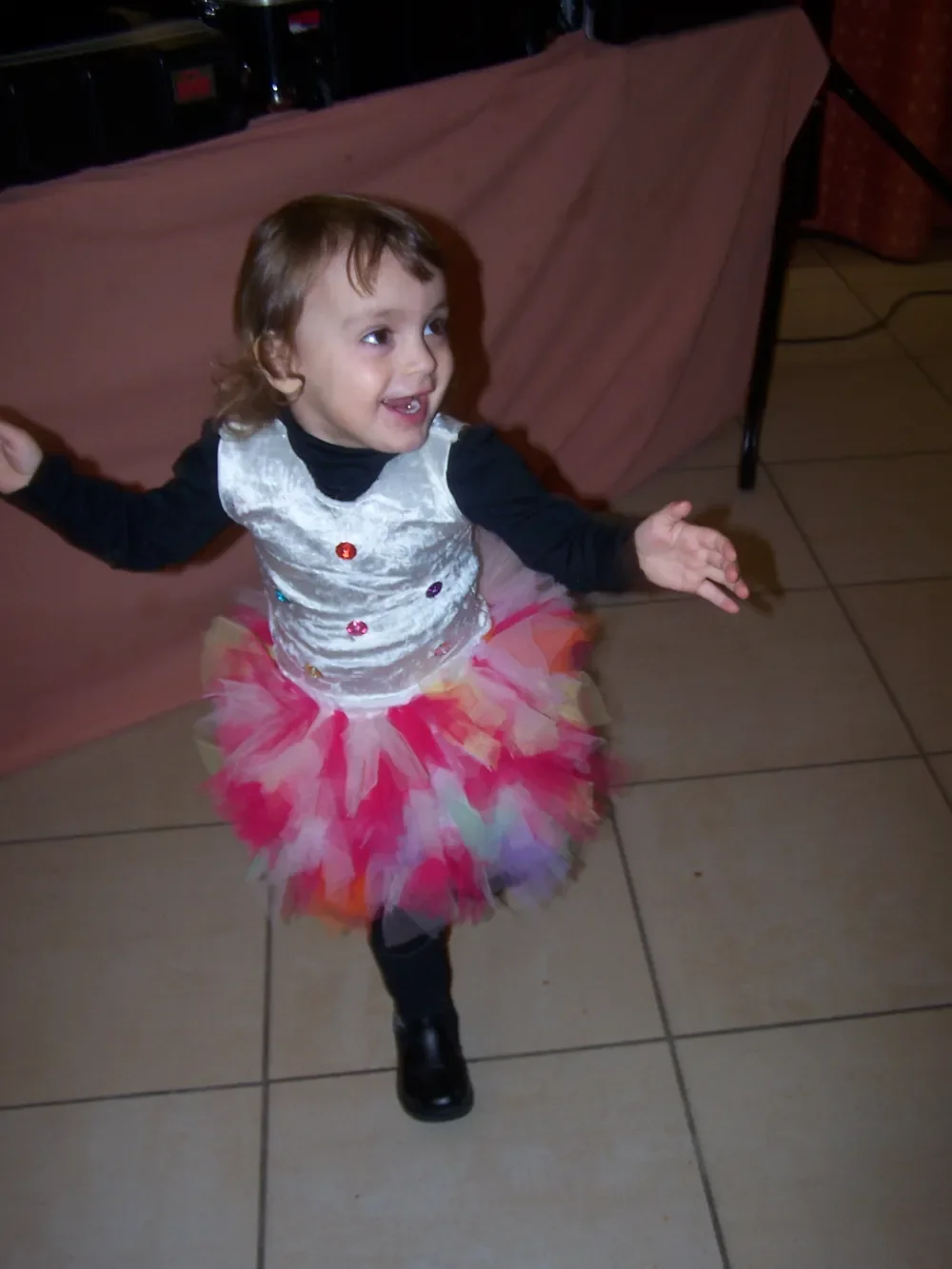 La même petite fille qui danse sur un carrelage, les bras écartés, dans sa robe au haut blanc brillant et à la jupe tutu en tulle rose, rouge, blanc et bleu pâle
