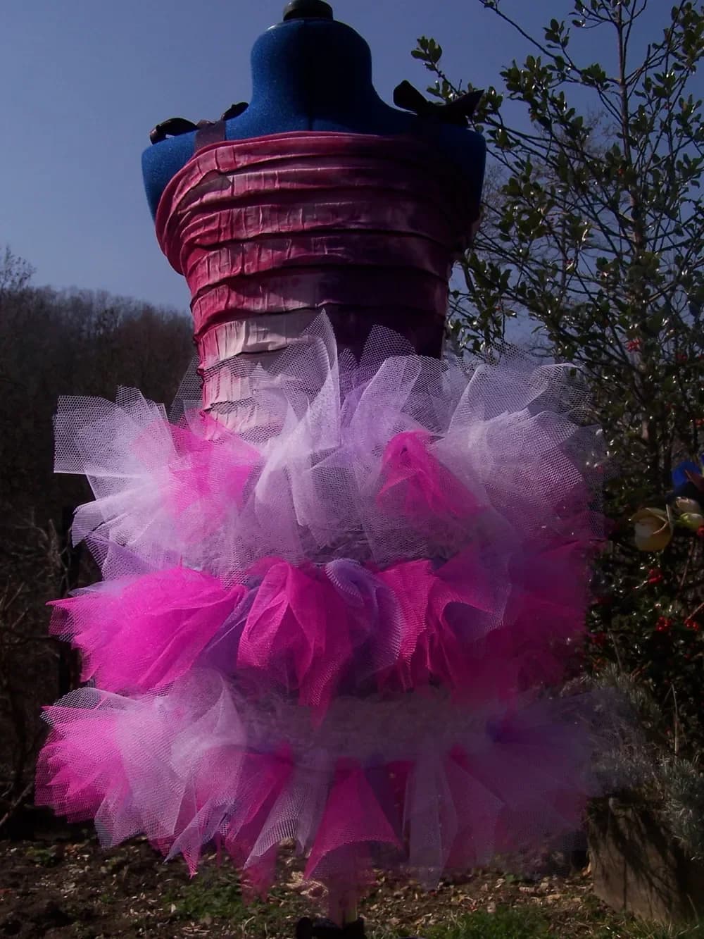 Robe bustier sur un buste de couture en extérieur, devant des arbres : haut en tissu plissé bordeaux et jupe tutu à plusieurs étages de tulle fuchsia, rose et blanc