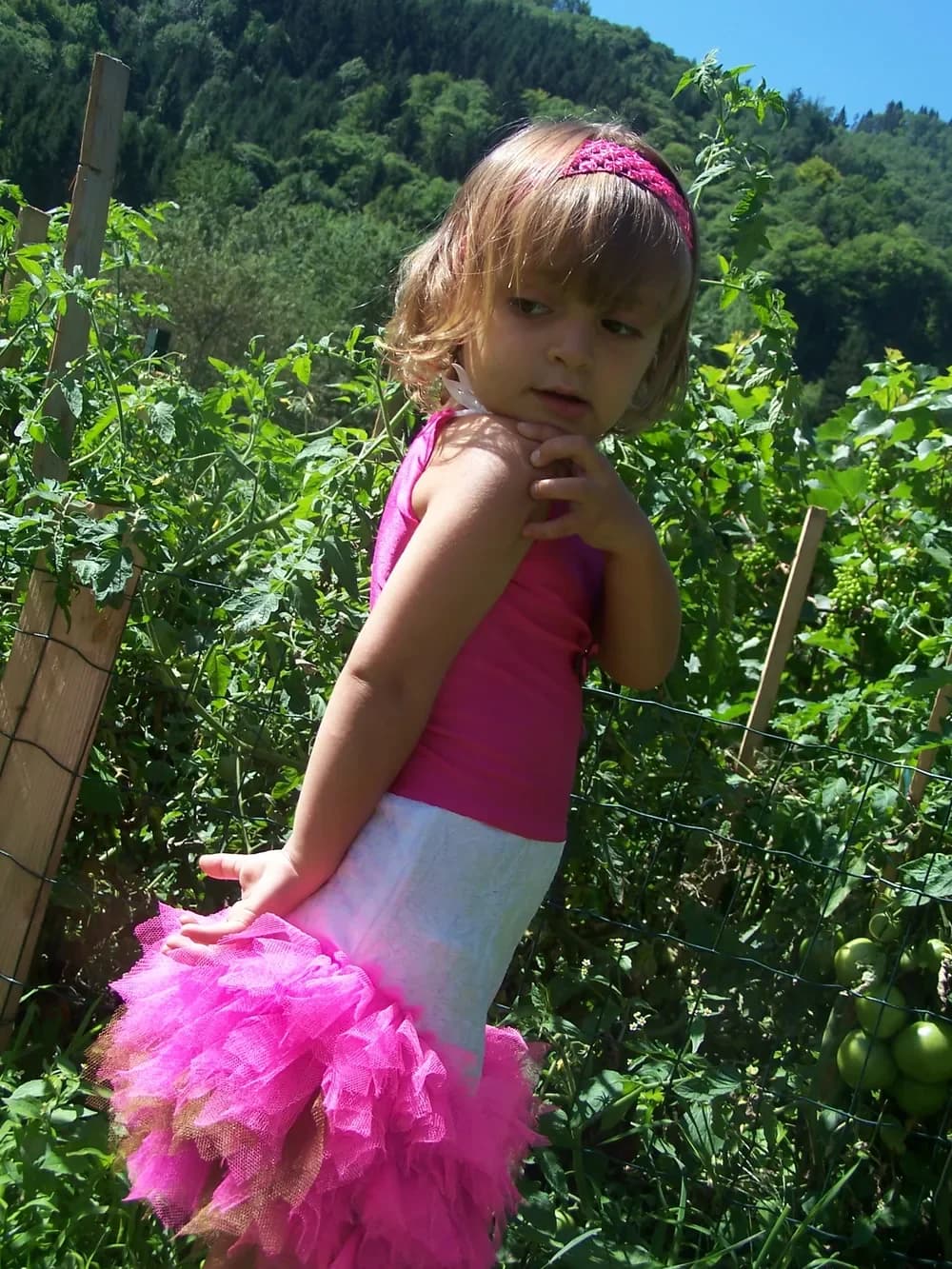 Petite fille vue de profil dans un potager, en robe au haut rose fuchsia, ceinture de dentelle blanche et jupe tutu en tulle rose fuchsia, un bandeau rose dans les cheveux