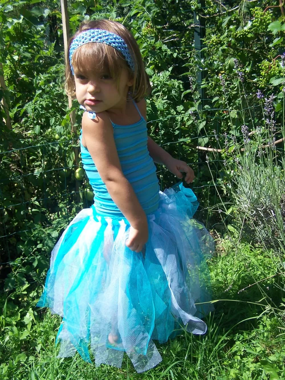 Petite fille dans un jardin, avec un bandeau turquoise, un débardeur rayé turquoise et une longue jupe tutu en tulle turquoise et blanc