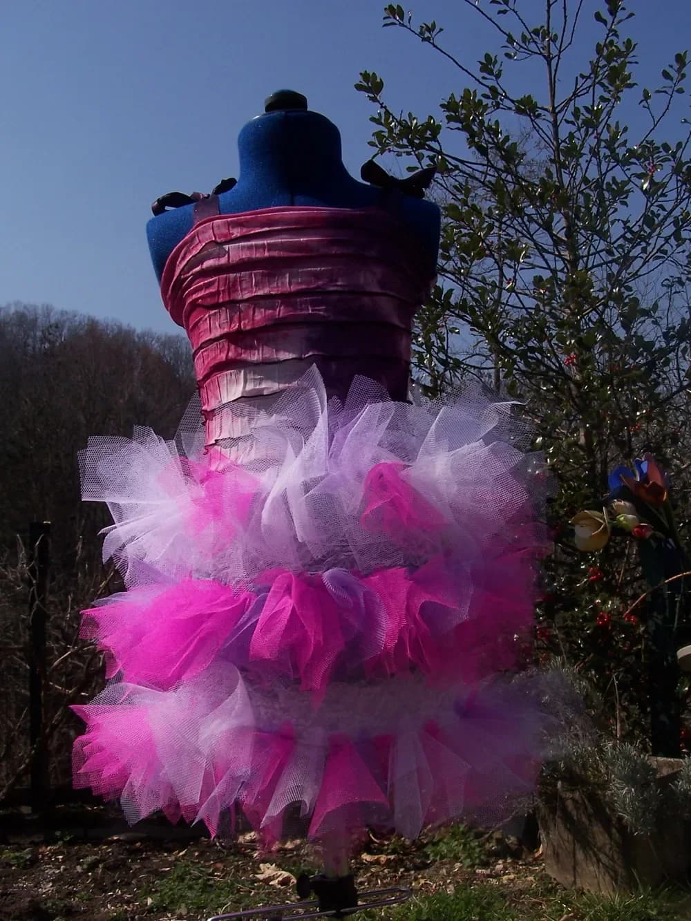 La robe au haut plissé bordeaux sur un buste de couture, vue sous un ciel bleu, avec sa jupe tutu à étages de tulle fuchsia, rose et blanc