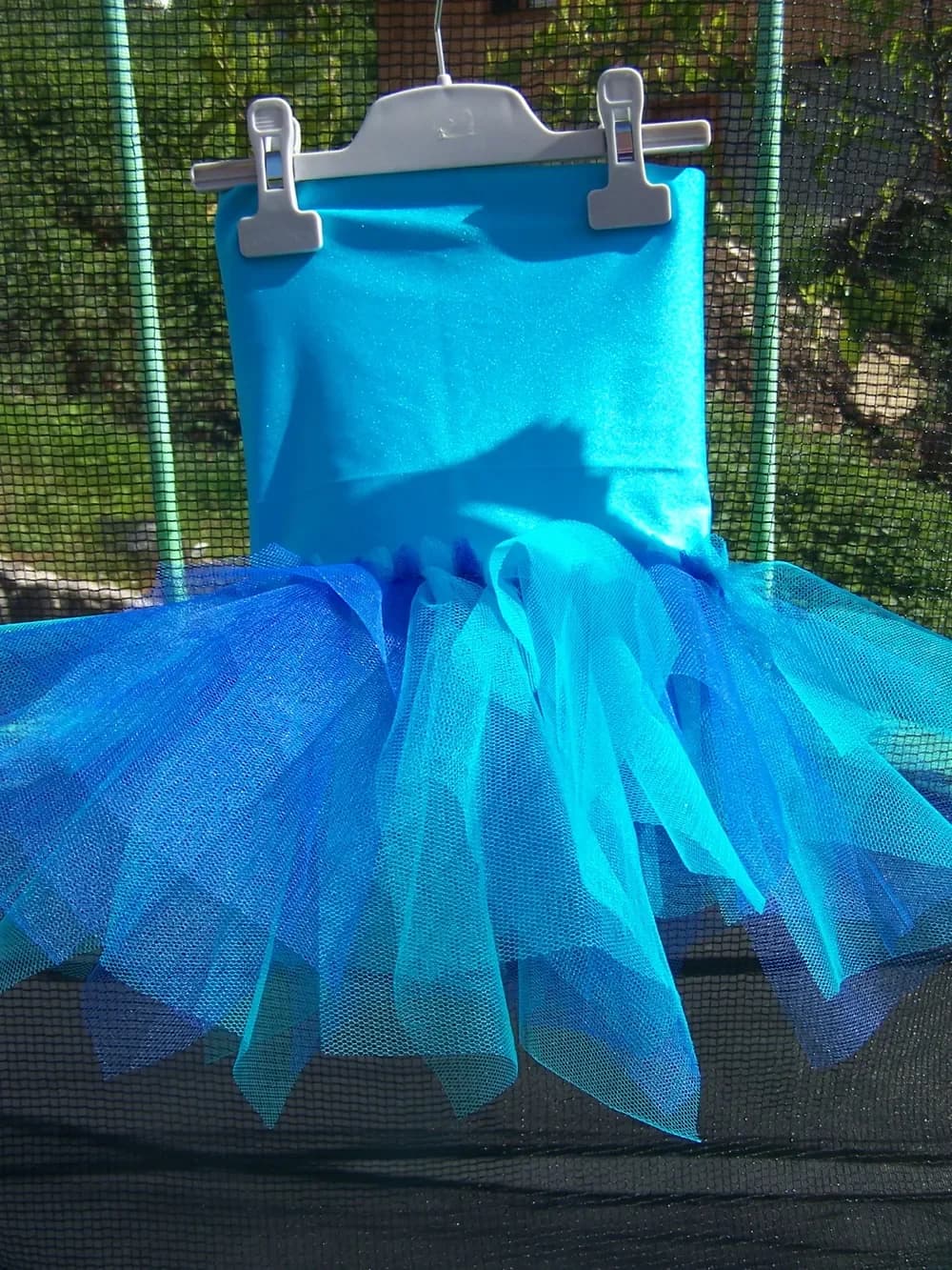 Robe suspendue à un cintre blanc devant un trampoline : haut bustier en jersey turquoise et jupe en tulle turquoise et bleu aux pointes irrégulières