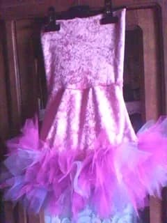 Robe sur un cintre devant une armoire en bois : haut en tissu rose à motifs damassés argentés et bas bordé de tulle rose, violet et blanc
