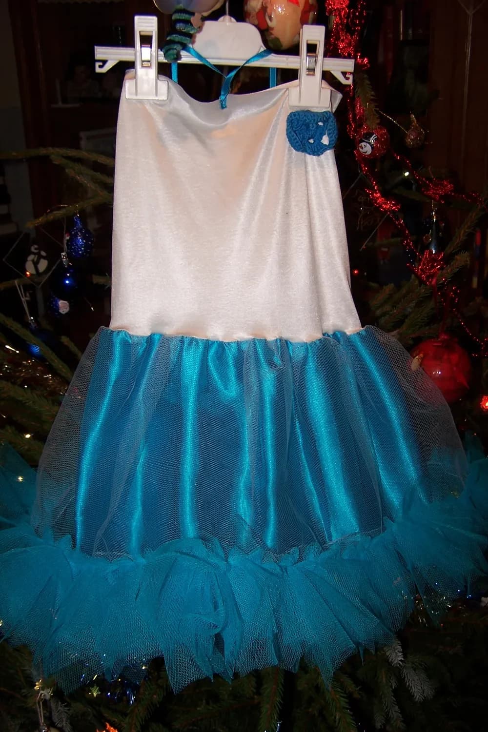 Robe sur un cintre devant un sapin de Noël : haut blanc satiné orné d'une petite fleur bleue et jupe en satin turquoise bordée d'un volant de tulle turquoise