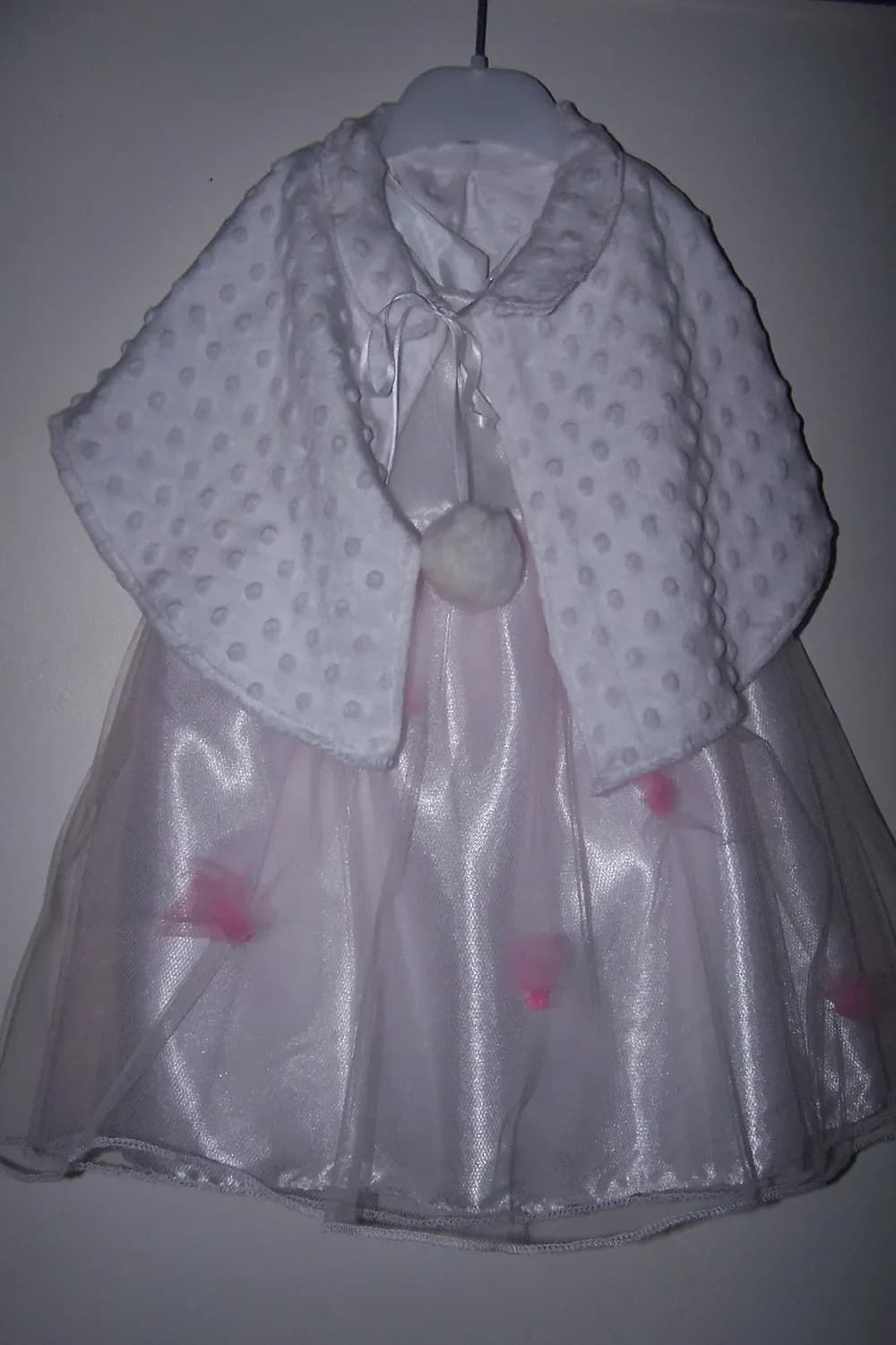 Robe blanche en satin sur un cintre, recouverte d'un petit boléro blanc en tissu à pois en relief noué au cou par des pompons, avec de petites roses roses sur la jupe