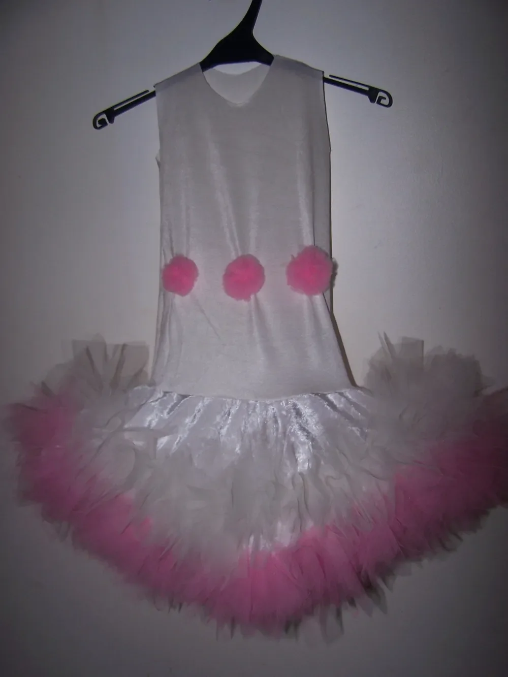 Robe blanche sans manches sur un cintre noir, avec trois pompons roses à la taille et une jupe en satin blanc bordée de volants de tulle rose