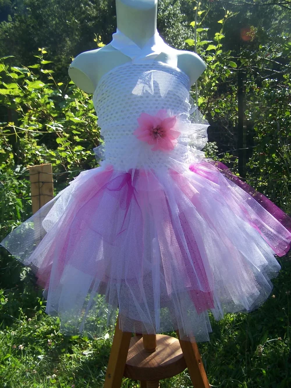 Robe sur un buste de couture posé sur un tabouret dans un jardin ensoleillé : haut en dentelle crochet blanche orné d'une fleur rose et jupe en tulle blanc et rose, avec quelques bandes violettes
