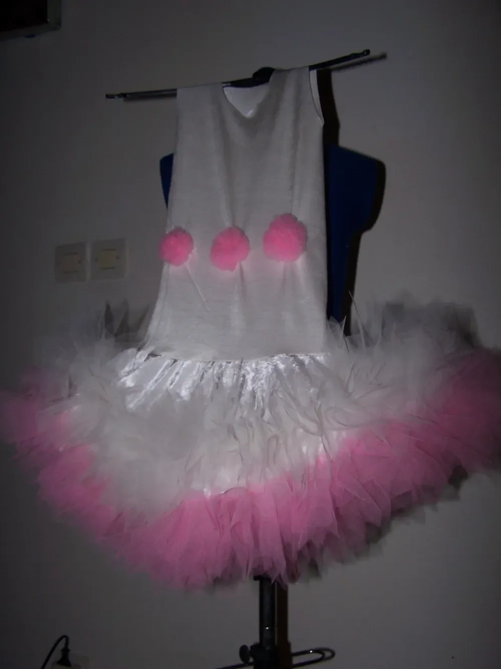 La robe blanche aux trois pompons roses présentée sur un buste dans une pièce, avec sa jupe très gonflante en tulle blanc et rose