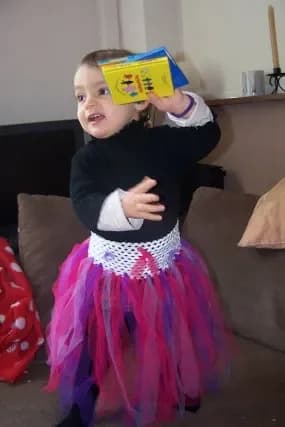 Petite fille dans un salon, qui tient une boîte jaune au-dessus de sa tête, vêtue d'un pull noir, d'une ceinture en dentelle crochet blanche et d'une jupe en longues bandes de tulle fuchsia et violet