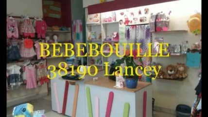 Intérieur de la boutique Bébébouille : un comptoir blanc décoré de bandes de couleur, des vêtements de bébé roses et blancs suspendus aux murs, des étagères garnies de peluches et d'accessoires, avec en surimpression en lettres jaunes « BEBEBOUILLE 38190 Lancey »