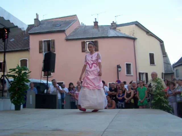 Megguy sur le podium du défilé de La Mure, en plein air, vêtue d'une robe de princesse au corsage rose orné de drapés et de rubans, et à la grande jupe bouffante blanche, devant des façades roses et un public nombreux massé derrière la scène
