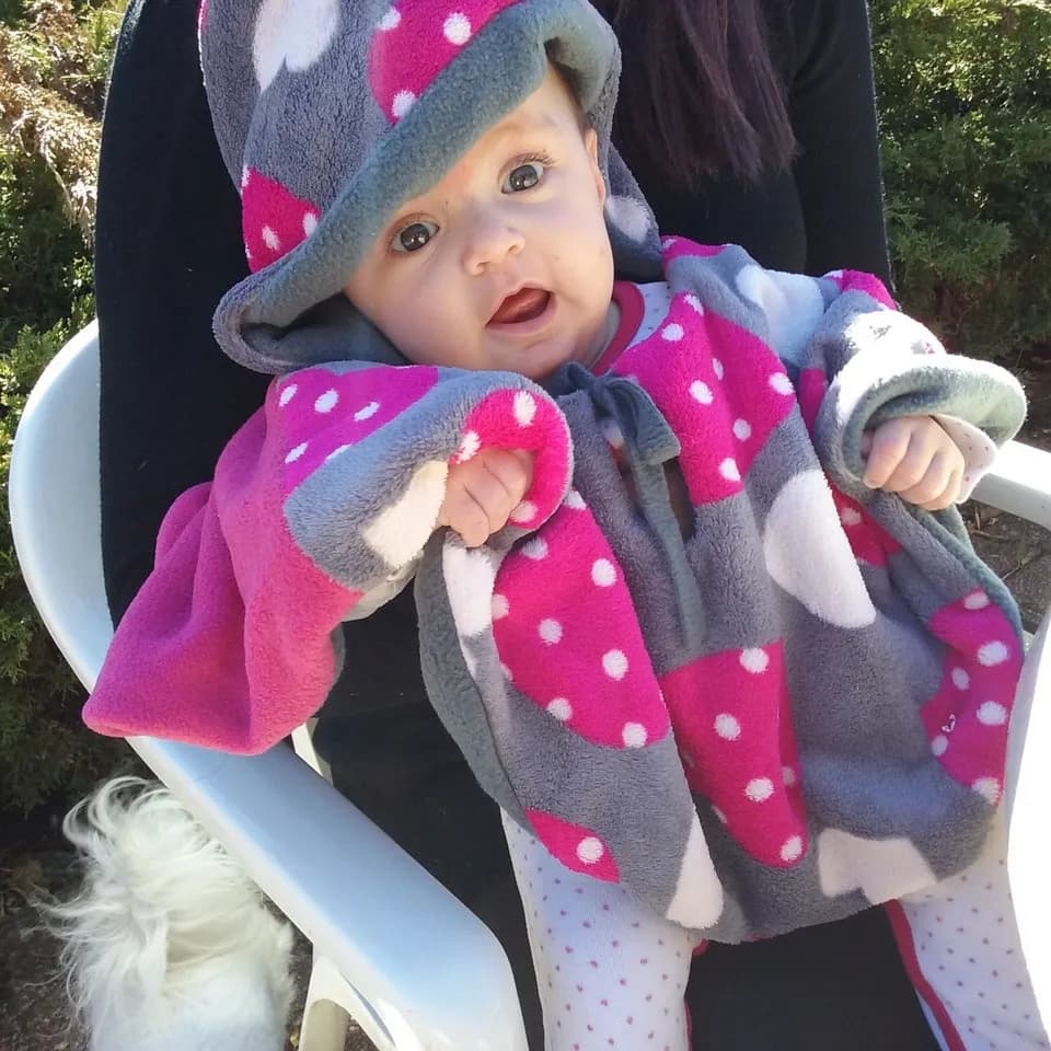 Bébé souriant emmitouflé dans une cape polaire réversible grise et rose à pois, avec capuche