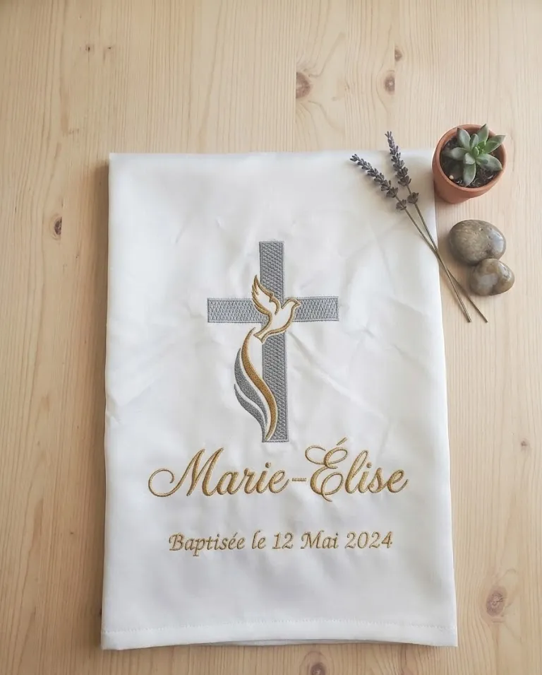Écharpe de baptême blanche brodée du prénom Marie-Élise, d'une croix et d'une colombe, posée sur une table en bois avec un brin de lavande