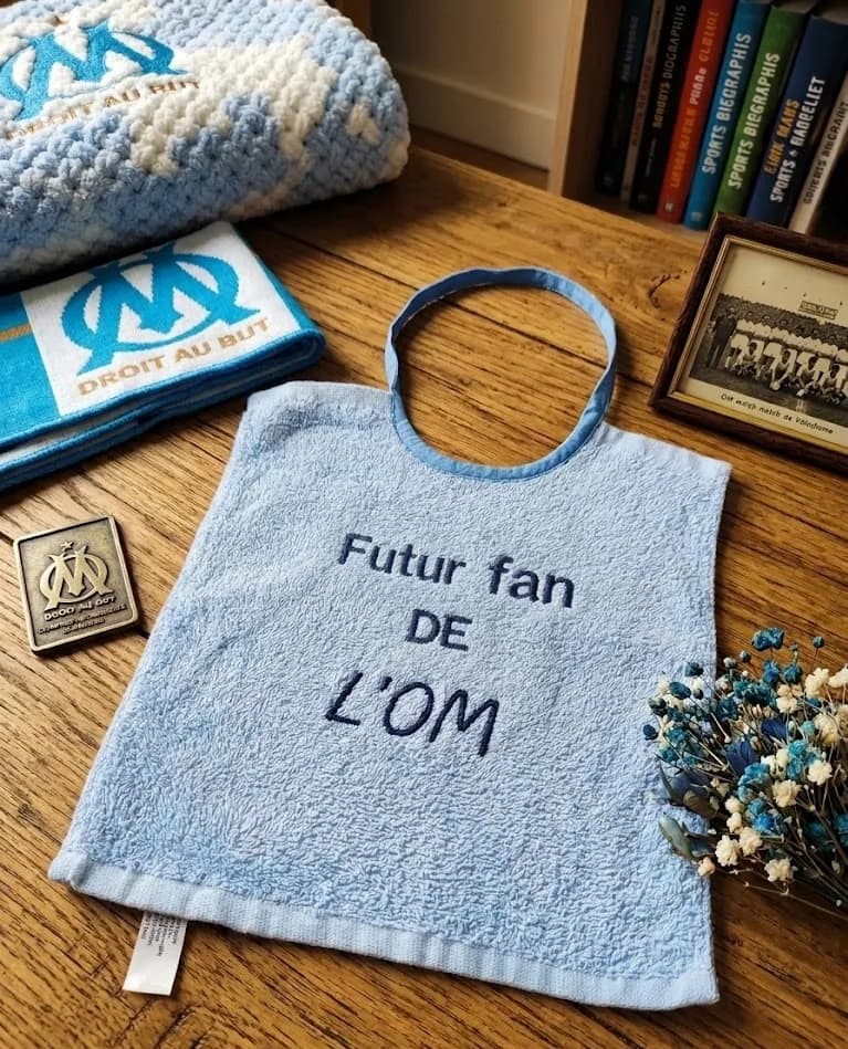 Bavoir en éponge bleu ciel brodé « Futur fan de l'OM », posé sur une table en bois avec des souvenirs du club de l'Olympique de Marseille