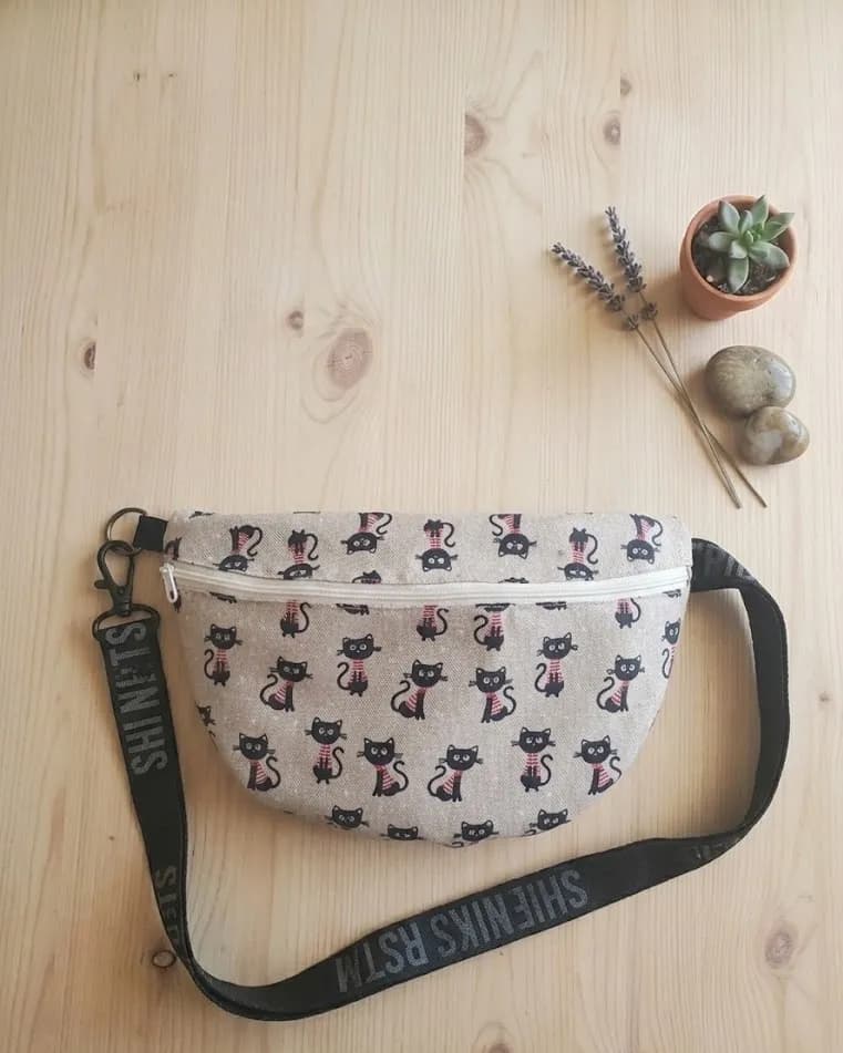 Sac banane en toile beige à motifs de chats noirs, avec bandoulière noire à inscription et pompon assorti, posé sur une table en bois clair